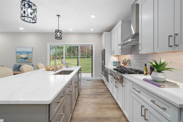 kitchen featuring a kitchen island with sink, stainless steel appliances, a sink, wall chimney exhaust hood, and light wood finished floors