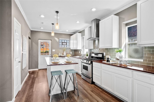 kitchen with wall chimney range hood, dark wood-type flooring, stainless steel gas range, a center island, and decorative light fixtures