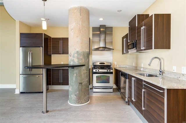 kitchen with appliances with stainless steel finishes, pendant lighting, sink, wall chimney range hood, and light hardwood / wood-style flooring