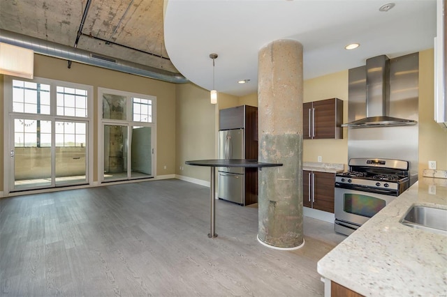 kitchen featuring appliances with stainless steel finishes, hanging light fixtures, light stone countertops, wall chimney range hood, and light hardwood / wood-style flooring