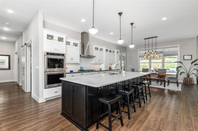 kitchen with white cabinets, wall chimney range hood, dark wood-type flooring, hanging light fixtures, and stainless steel appliances