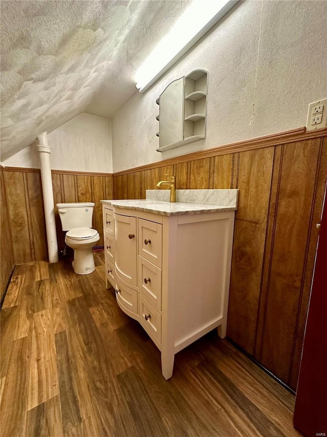 half bath with a textured ceiling, lofted ceiling, toilet, a wainscoted wall, and wood finished floors