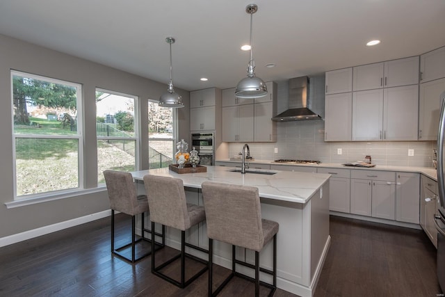 kitchen with sink, wall chimney exhaust hood, dark wood-type flooring, light stone counters, and a center island with sink