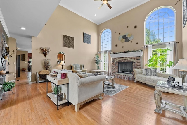 living room with a high ceiling, ornamental molding, a brick fireplace, ceiling fan, and light wood-type flooring