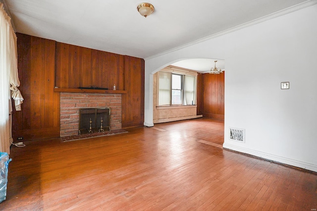 unfurnished living room featuring baseboard heating, hardwood / wood-style floors, wooden walls, ornamental molding, and a fireplace