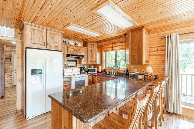 kitchen featuring light wood-type flooring, wooden walls, a kitchen bar, kitchen peninsula, and white appliances