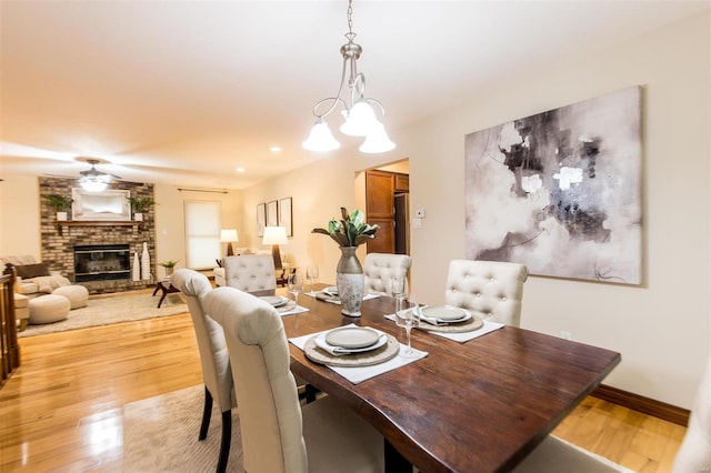 dining room with a brick fireplace, light wood-type flooring, and ceiling fan with notable chandelier