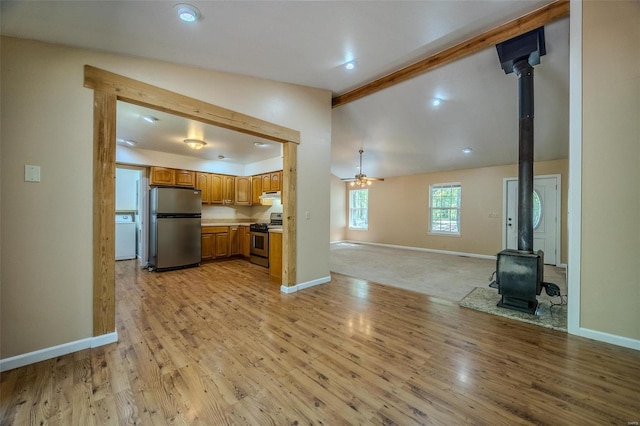 unfurnished living room featuring a wood stove, ceiling fan, light wood-type flooring, and lofted ceiling with beams