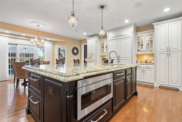 kitchen with stainless steel microwave, decorative light fixtures, a kitchen island with sink, white cabinets, and light hardwood / wood-style flooring