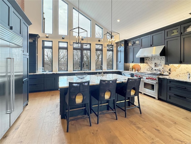 kitchen with high end appliances, light wood-type flooring, a center island with sink, and light stone countertops