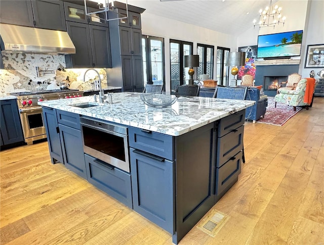 kitchen with light hardwood / wood-style flooring, appliances with stainless steel finishes, a chandelier, decorative backsplash, and a kitchen island with sink