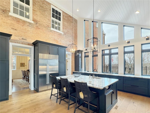 kitchen with built in fridge, light wood-type flooring, a towering ceiling, and an island with sink