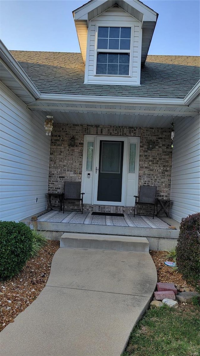 doorway to property featuring a porch, roof with shingles, and brick siding