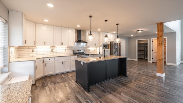 kitchen with appliances with stainless steel finishes, white cabinetry, a kitchen island with sink, light stone counters, and wall chimney range hood