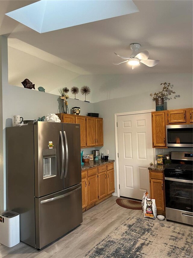 kitchen featuring ceiling fan, appliances with stainless steel finishes, lofted ceiling with skylight, dark stone countertops, and light hardwood / wood-style floors