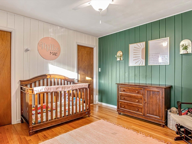 bedroom featuring a crib, light wood-type flooring, wood walls, and ceiling fan