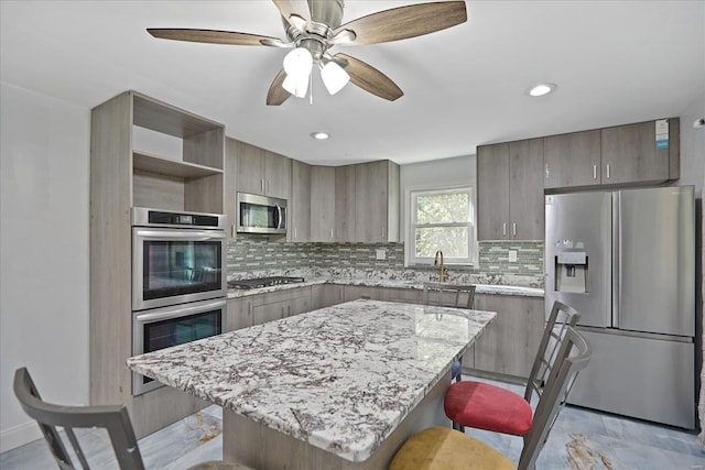 kitchen with light stone counters, backsplash, a kitchen island, stainless steel appliances, and ceiling fan