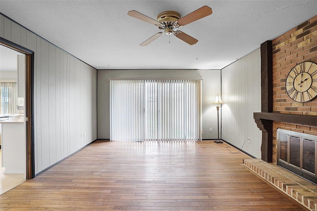 unfurnished living room featuring a brick fireplace, a textured ceiling, ceiling fan, light hardwood / wood-style flooring, and wood walls