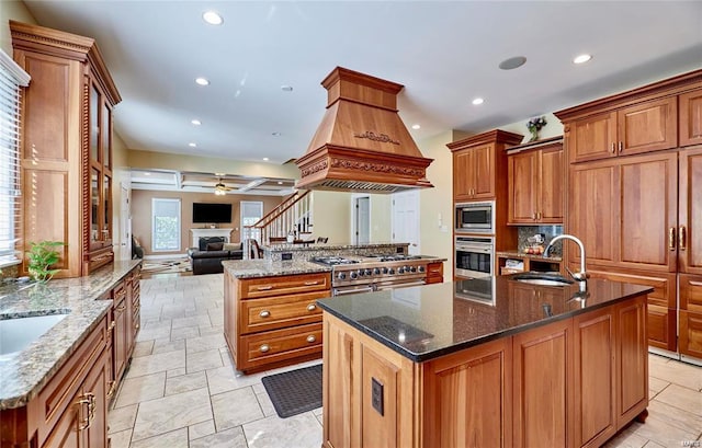 kitchen featuring dark stone counters, sink, custom exhaust hood, stainless steel appliances, and a center island with sink