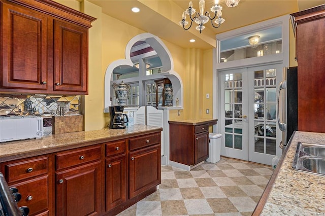 kitchen with decorative backsplash, stainless steel refrigerator, light stone countertops, an inviting chandelier, and french doors