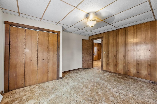 unfurnished bedroom with a drop ceiling, wooden walls, light carpet, and a closet