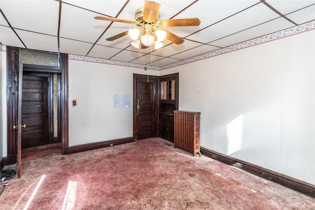 carpeted empty room with a drop ceiling, wooden walls, and ceiling fan