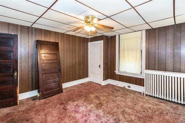 unfurnished bedroom featuring ceiling fan, a paneled ceiling, carpet flooring, radiator, and wooden walls