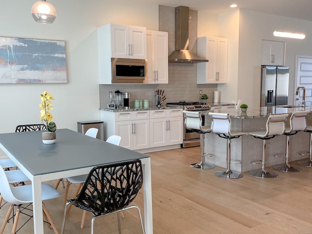 kitchen featuring appliances with stainless steel finishes, white cabinetry, light stone countertops, light hardwood / wood-style flooring, and wall chimney range hood