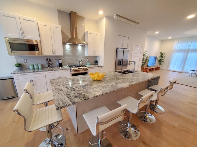 kitchen with sink, wall chimney exhaust hood, a kitchen island with sink, white cabinetry, and appliances with stainless steel finishes