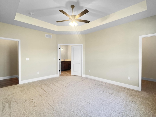 unfurnished bedroom with light carpet, a tray ceiling, ceiling fan, and ensuite bath