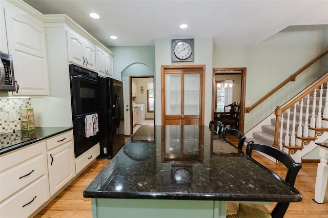 kitchen with white cabinets, a kitchen island with sink, black appliances, dark stone counters, and light hardwood / wood-style floors