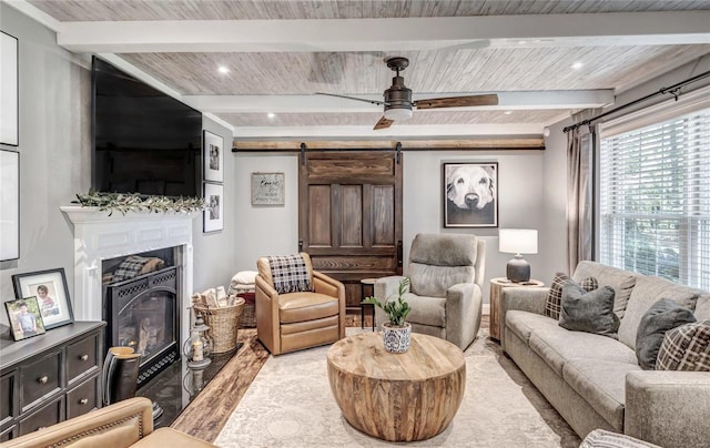 living area with a barn door, a glass covered fireplace, ceiling fan, wooden ceiling, and beamed ceiling