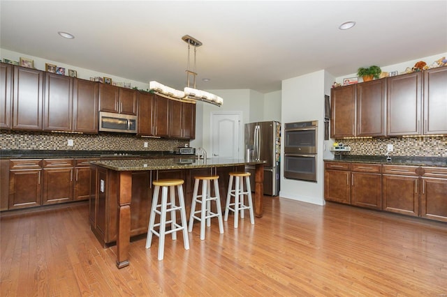 kitchen featuring appliances with stainless steel finishes, decorative light fixtures, dark stone countertops, a kitchen island with sink, and light wood-type flooring