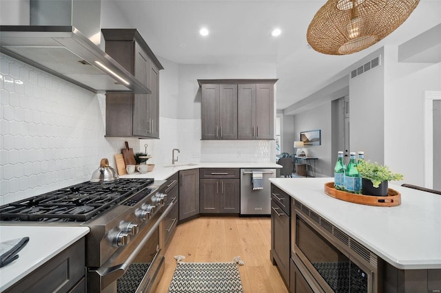 kitchen featuring pendant lighting, tasteful backsplash, wall chimney range hood, appliances with stainless steel finishes, and light wood-type flooring