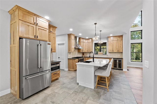 kitchen featuring an island with sink, stainless steel appliances, beverage cooler, wall chimney range hood, and hanging light fixtures