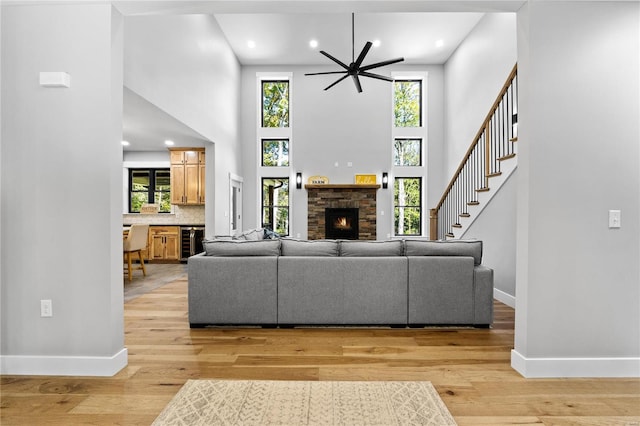 living room featuring ceiling fan, wine cooler, a stone fireplace, a high ceiling, and light wood-type flooring