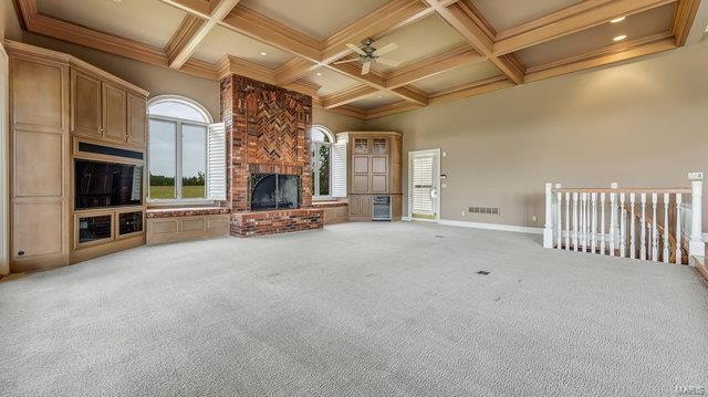 unfurnished living room featuring ceiling fan, beam ceiling, a brick fireplace, coffered ceiling, and carpet floors