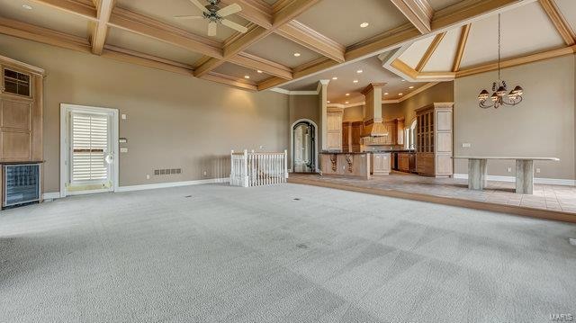 unfurnished living room with ceiling fan with notable chandelier, crown molding, coffered ceiling, beam ceiling, and light colored carpet