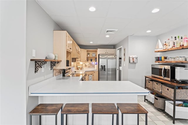 kitchen with stainless steel appliances, kitchen peninsula, a kitchen bar, light brown cabinetry, and light tile patterned floors