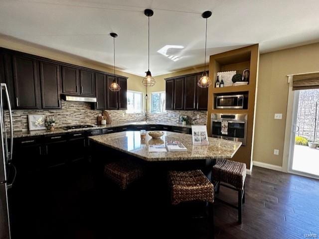 kitchen with a breakfast bar, under cabinet range hood, light stone counters, stainless steel appliances, and decorative backsplash