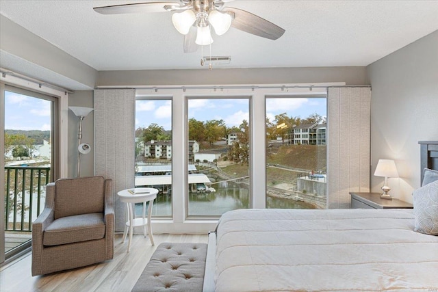 bedroom featuring access to outside, light wood-type flooring, a textured ceiling, a water view, and ceiling fan