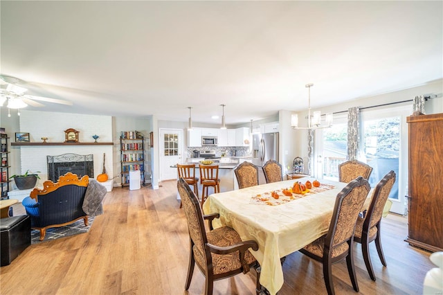 dining area featuring a brick fireplace, ceiling fan with notable chandelier, and light wood-type flooring