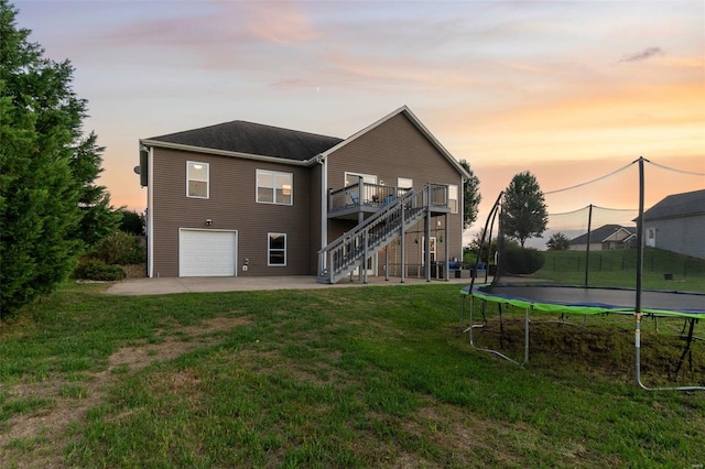 back house at dusk featuring a trampoline, a garage, a wooden deck, and a lawn