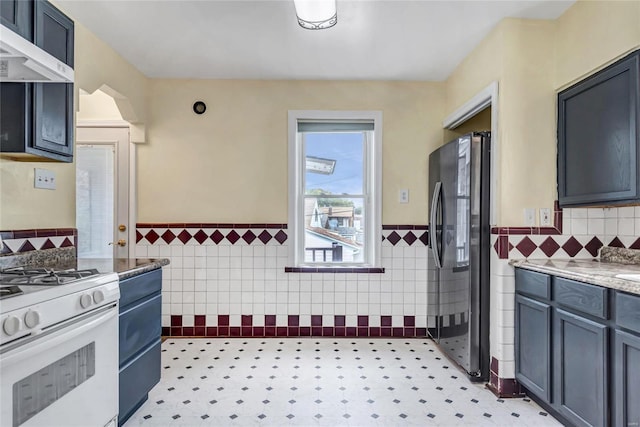 kitchen with white gas range, tile walls, light stone counters, extractor fan, and stainless steel refrigerator