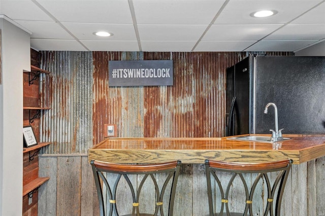 bar featuring sink, a drop ceiling, wooden walls, and black fridge