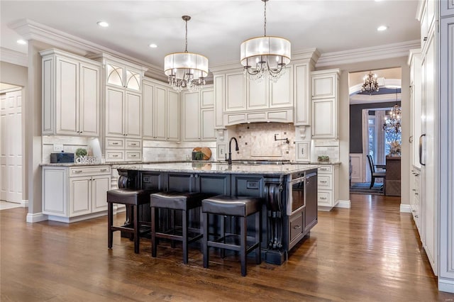 kitchen with an island with sink, ornamental molding, dark wood-style floors, an inviting chandelier, and light stone countertops