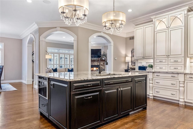 kitchen with a sink, crown molding, an inviting chandelier, arched walkways, and a kitchen island with sink