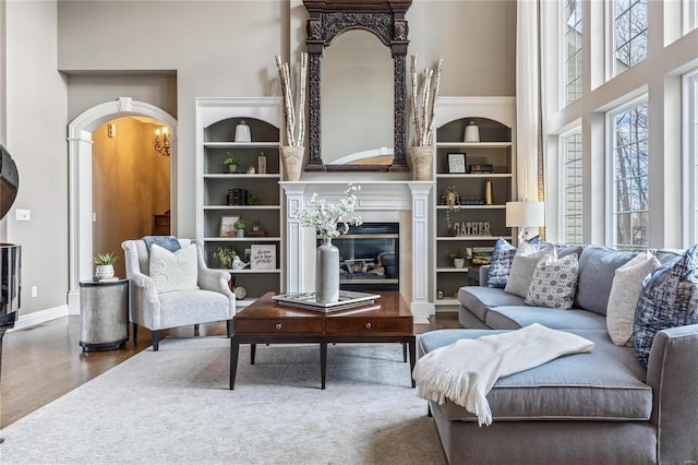 living area featuring built in shelves, wood finished floors, a glass covered fireplace, baseboards, and a towering ceiling