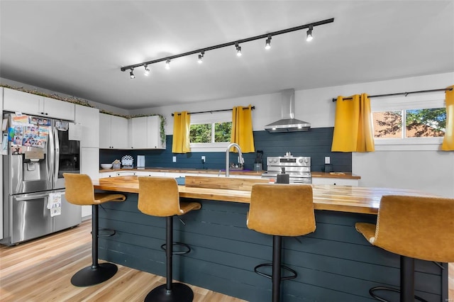 kitchen with wall chimney exhaust hood, butcher block countertops, light hardwood / wood-style floors, white cabinetry, and stainless steel appliances