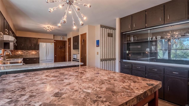 kitchen with tasteful backsplash, dark brown cabinets, an inviting chandelier, white fridge, and sink